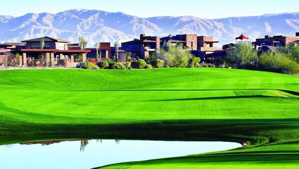 A golf course with a pond, surrounded by modern buildings with mountain views.