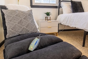 A massage chair with a control panel, a bedside table with a plant, and a bed with white bedding.