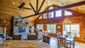 Un intérieur spacieux de chalet en bois, avec une cheminée en pierre, un coin repas et une grande fenêtre donnant sur l’extérieur.