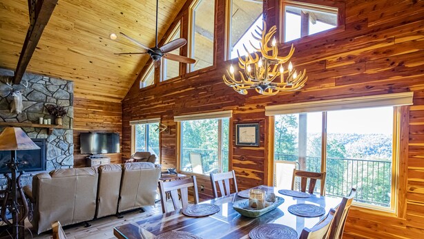 L’intérieur chaleureux d’un chalet avec une cheminée en pierre, une table à manger en bois et de grandes fenêtres offrant une vue panoramique.
