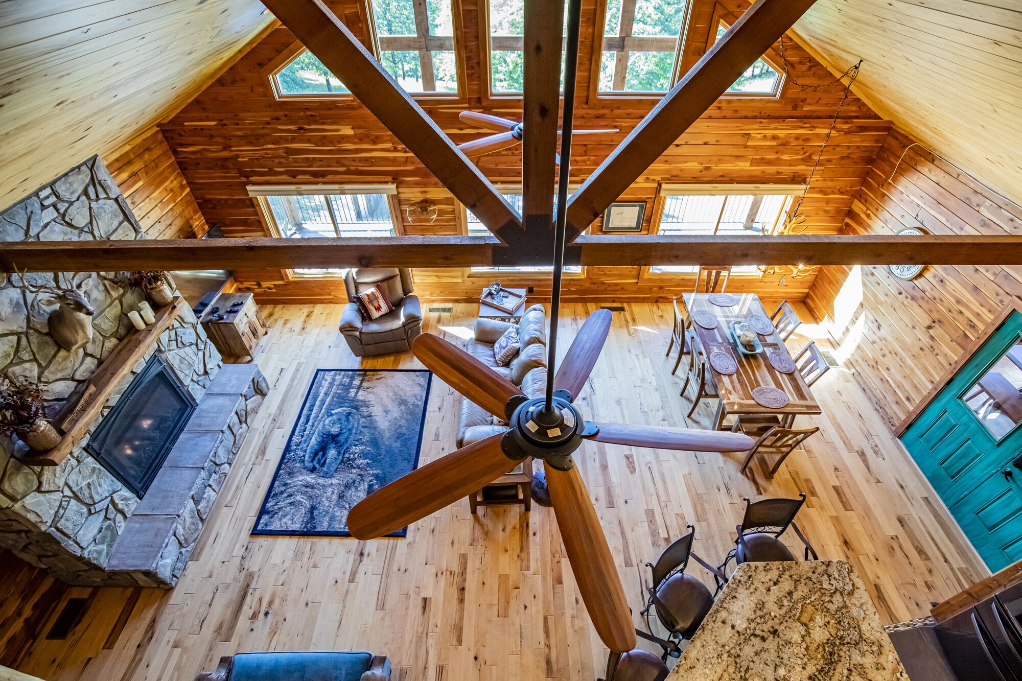 Une cabane en bois aux plafonds hauts, dotée d’un ventilateur de plafond central, d’une cheminée en pierre et d’un coin repas avec une table et des chaises.
