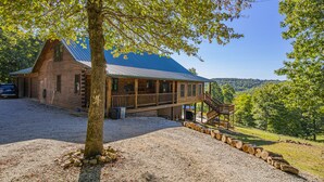 Un chalet rustique doté d’un porche et d’un escalier menant à une terrasse, entouré d’une allée en gravier et d’un paysage boisé.
