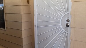 A white metal door with a radial mesh pattern, a small window with blinds, and a doorbell on the wall.