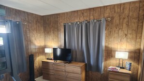 A room with wooden panel walls, a TV on a wooden dresser, two lamps, and a small table with books and a box.