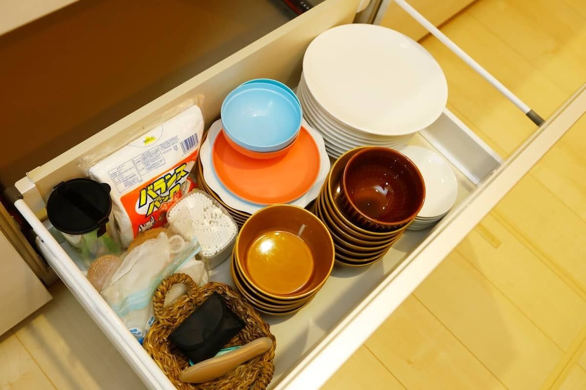 A kitchen drawer with various items including plates, bowls, a basket, and a package of food.