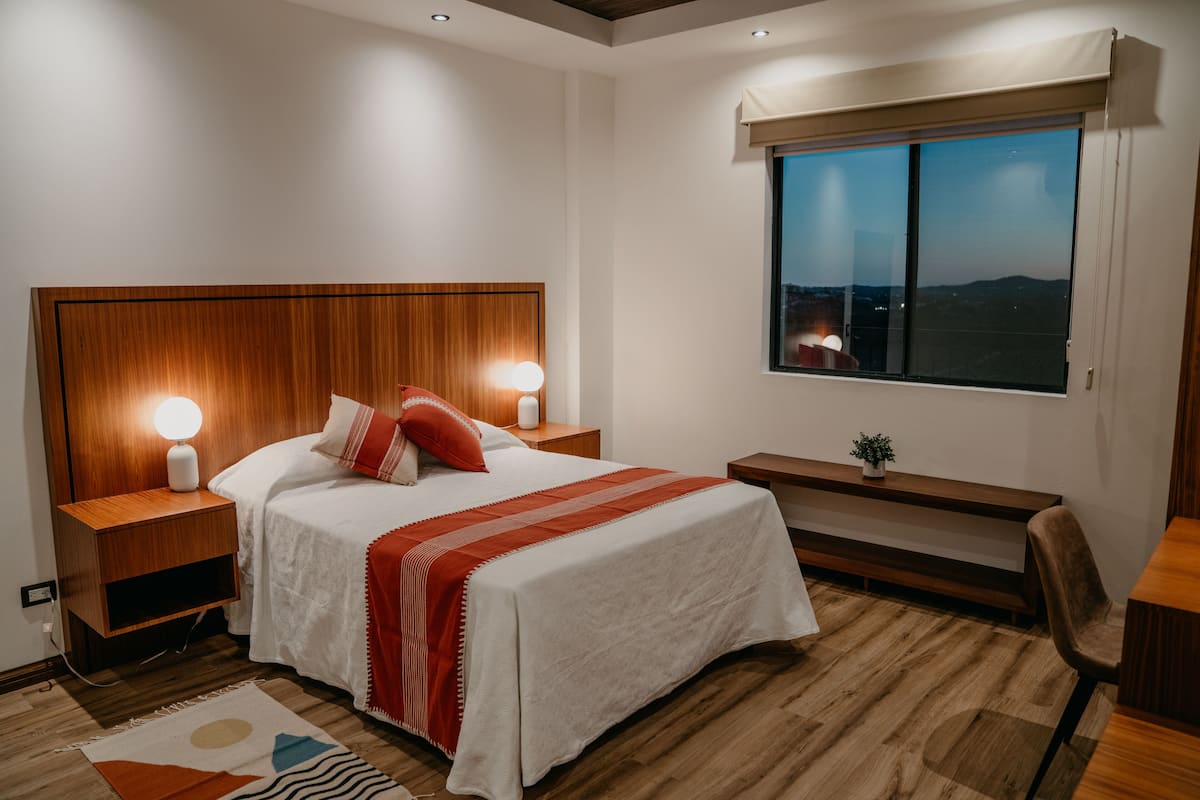 Ein Schlafzimmer mit Holz-Kopfteil, einem großen Bett mit weißer Bettwäsche und roten Akzenten, Nachttischlampen, einer Bank und Blick auf die Stadt im Abendrot.