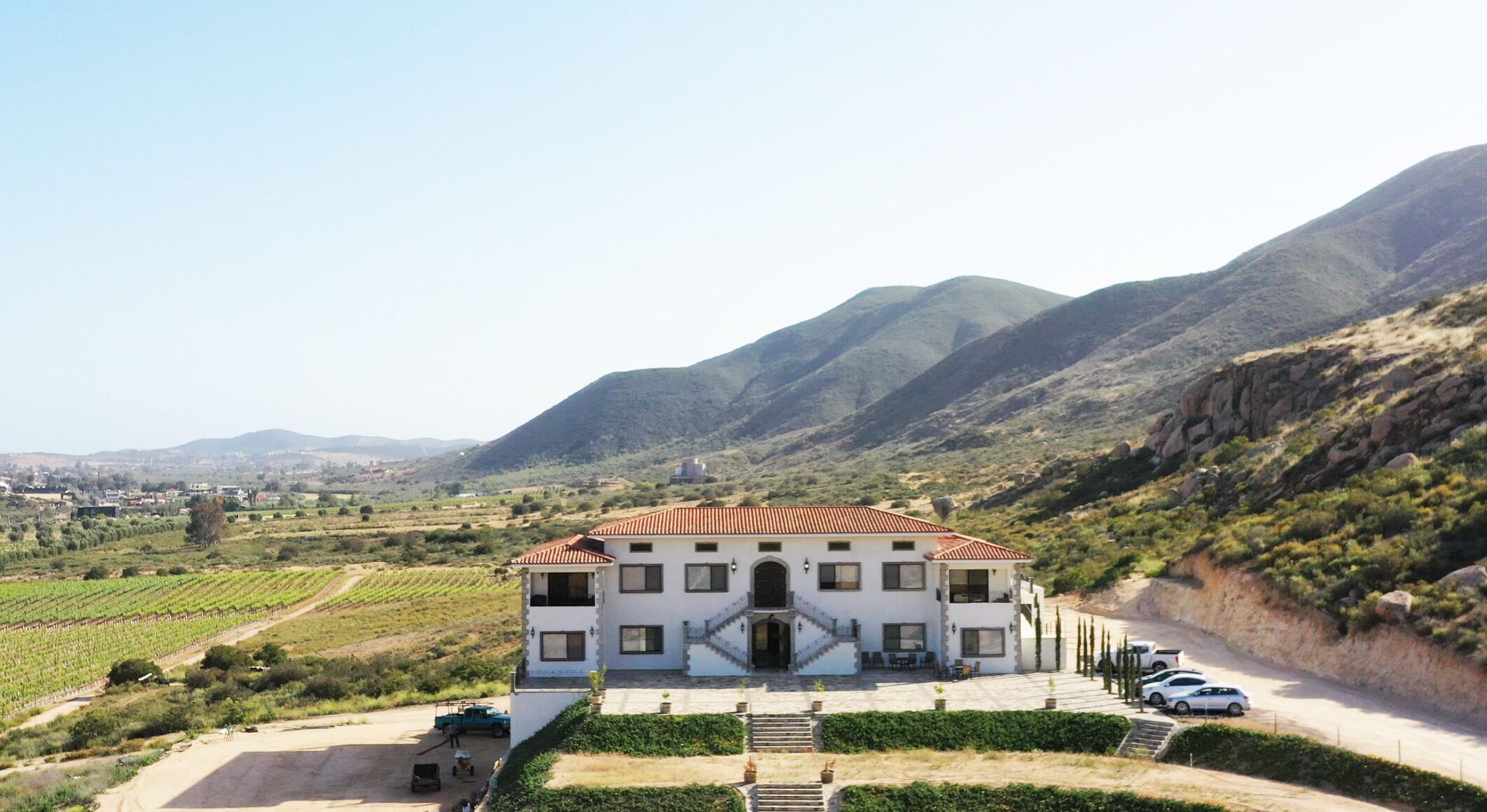 A large white building with a red-tiled roof, surrounded by vineyards and mountains.