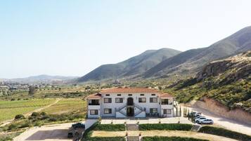 A large white building with a red-tiled roof, surrounded by vineyards and mountains.