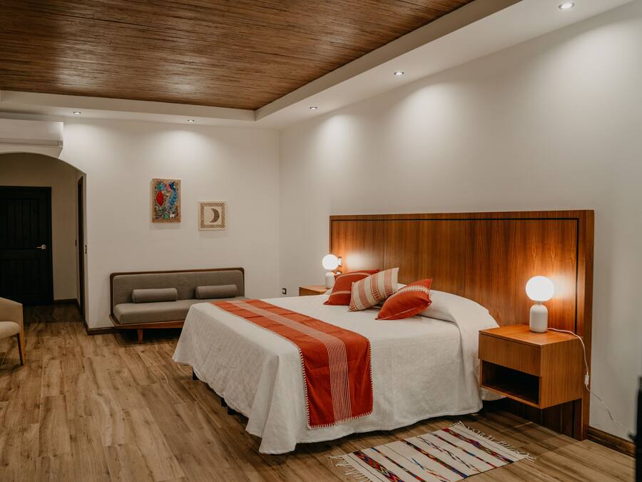 A modern bedroom with a wooden ceiling, a large bed with white bedding and red accents, a bedside table with a lamp, a sofa, and a small table.