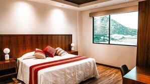 A modern bedroom with a wooden headboard, a large window with a mountain view, a bedside table with a lamp, and a patterned rug. -  Villa Montefiori (Valle de Guadalupe)