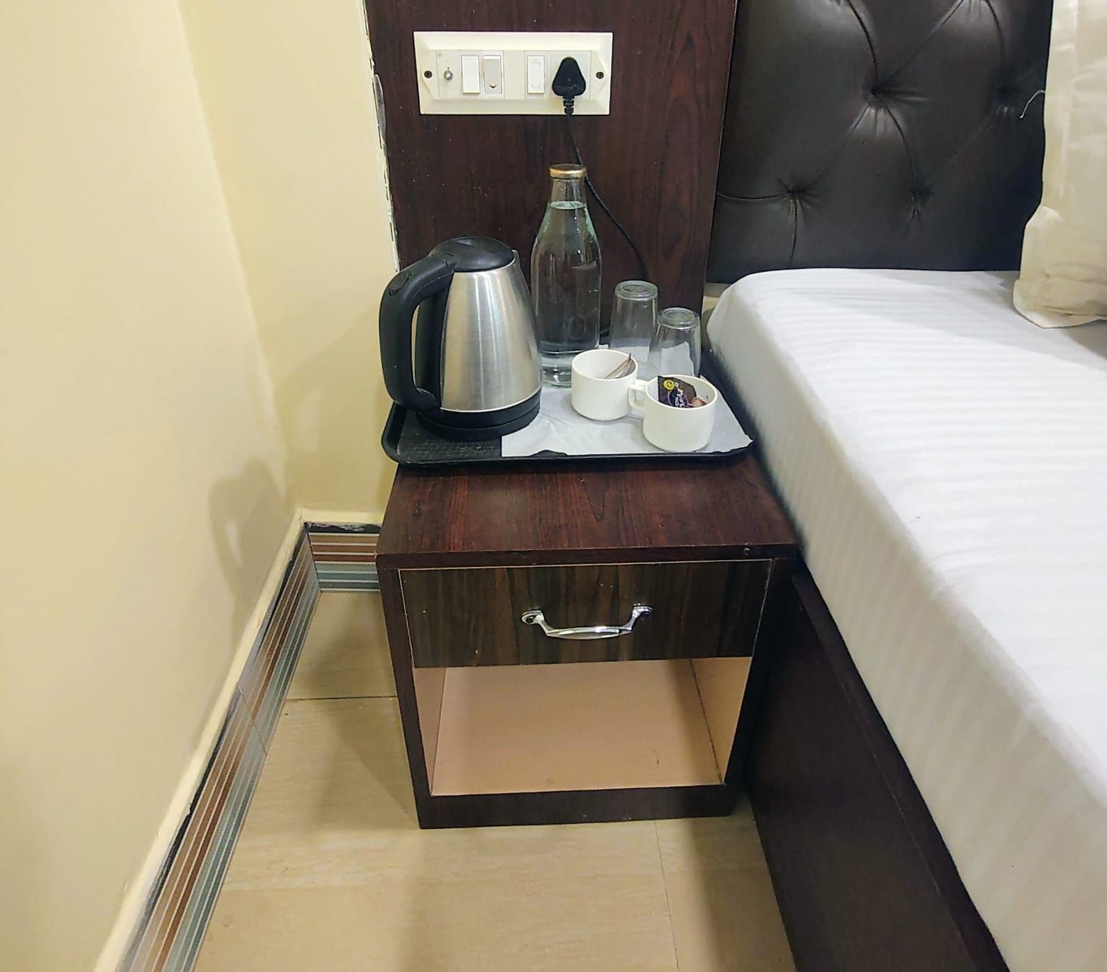 A hotel room with a bed, a nightstand with a kettle, cups, and glasses, and a drawer.