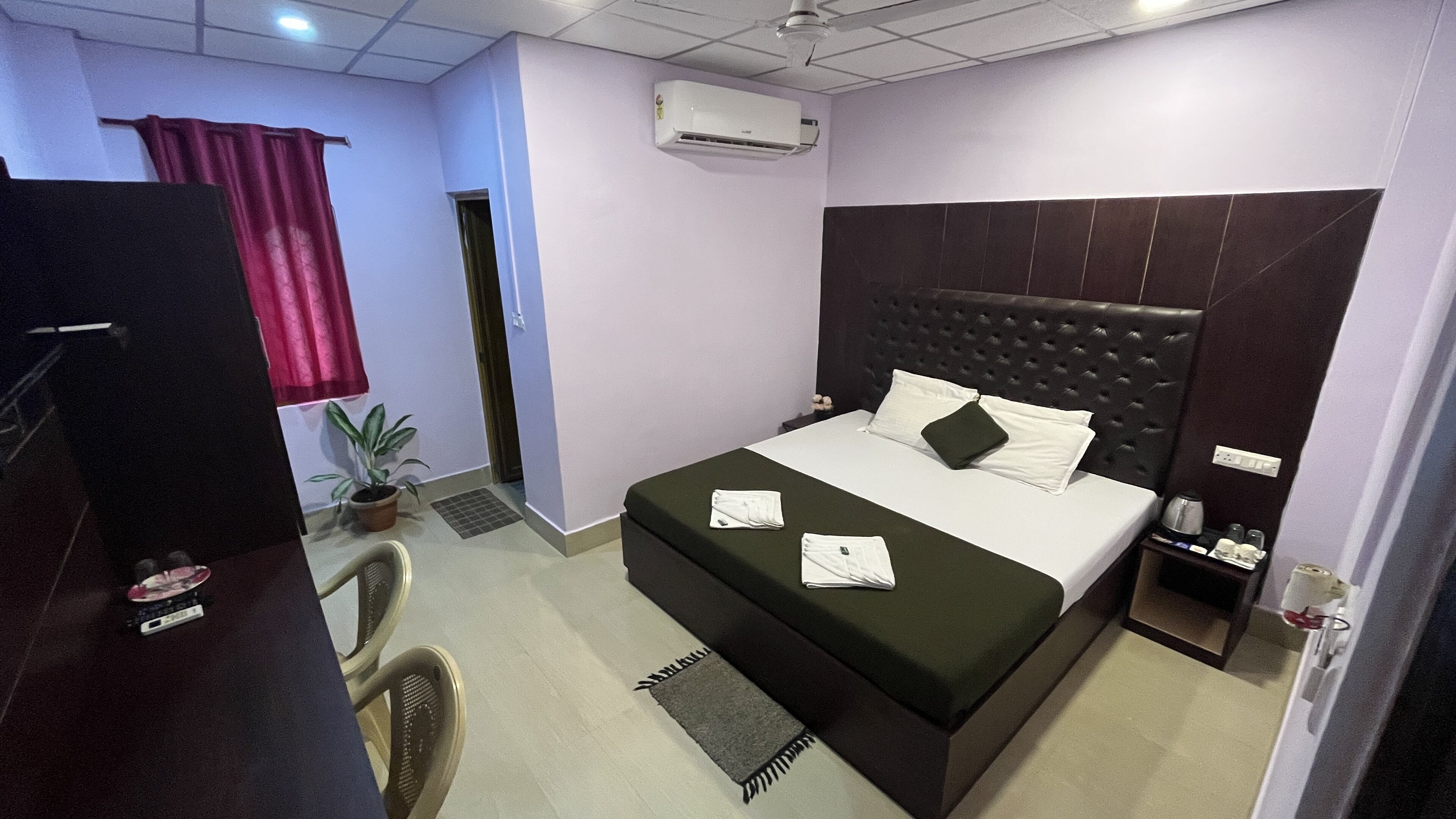 A hotel room with a large bed, a bedside table with a lamp, a potted plant, and a window with curtains.