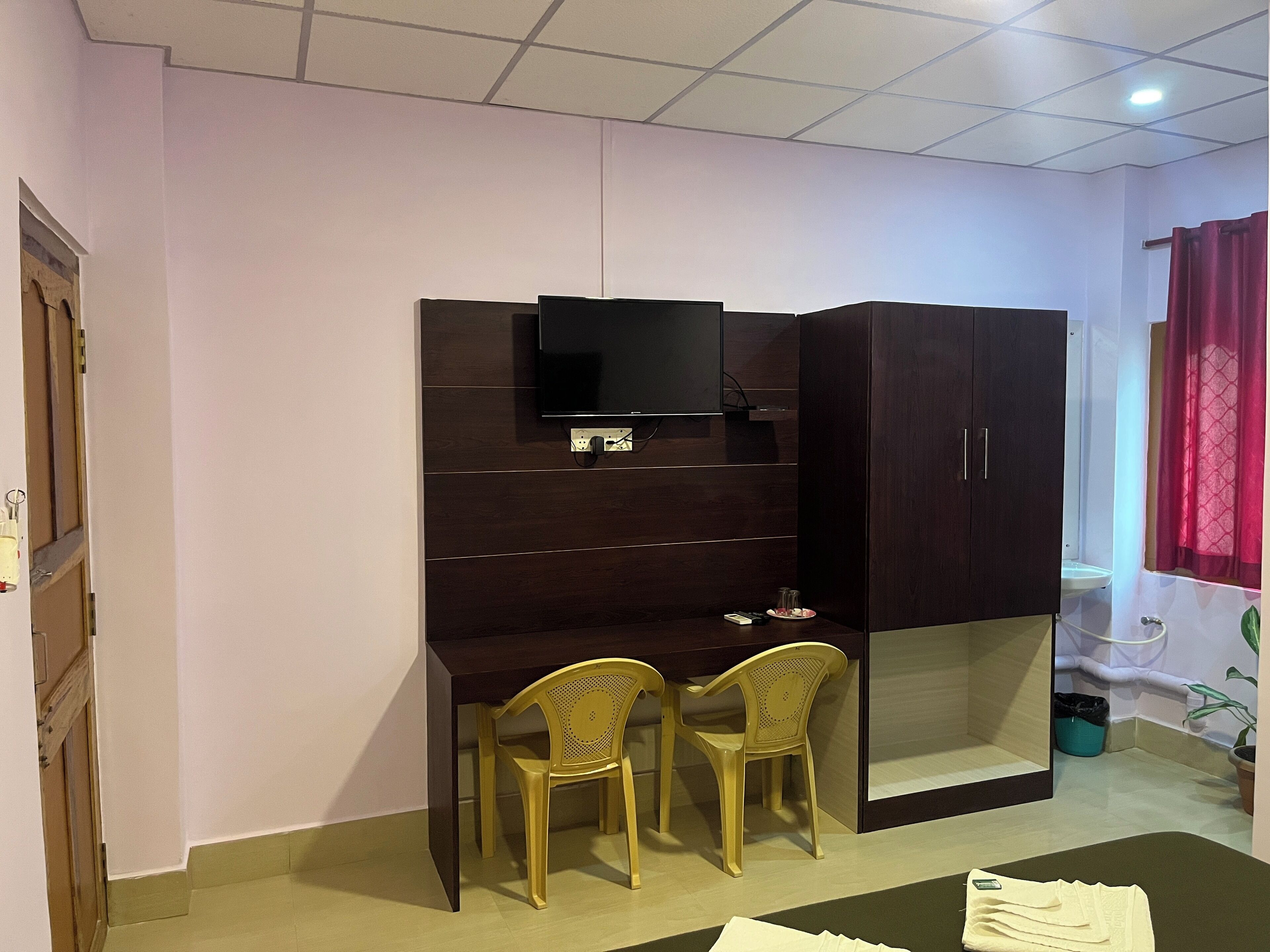 A room with a dark wooden wardrobe, a desk with two chairs, a television mounted on the wall, and a window with a red curtain.