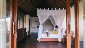 Ein Zimmer mit einem Bett unter einem Baldachin, Holzboden, einem kleinen Nachttisch und Blick ins Freie.