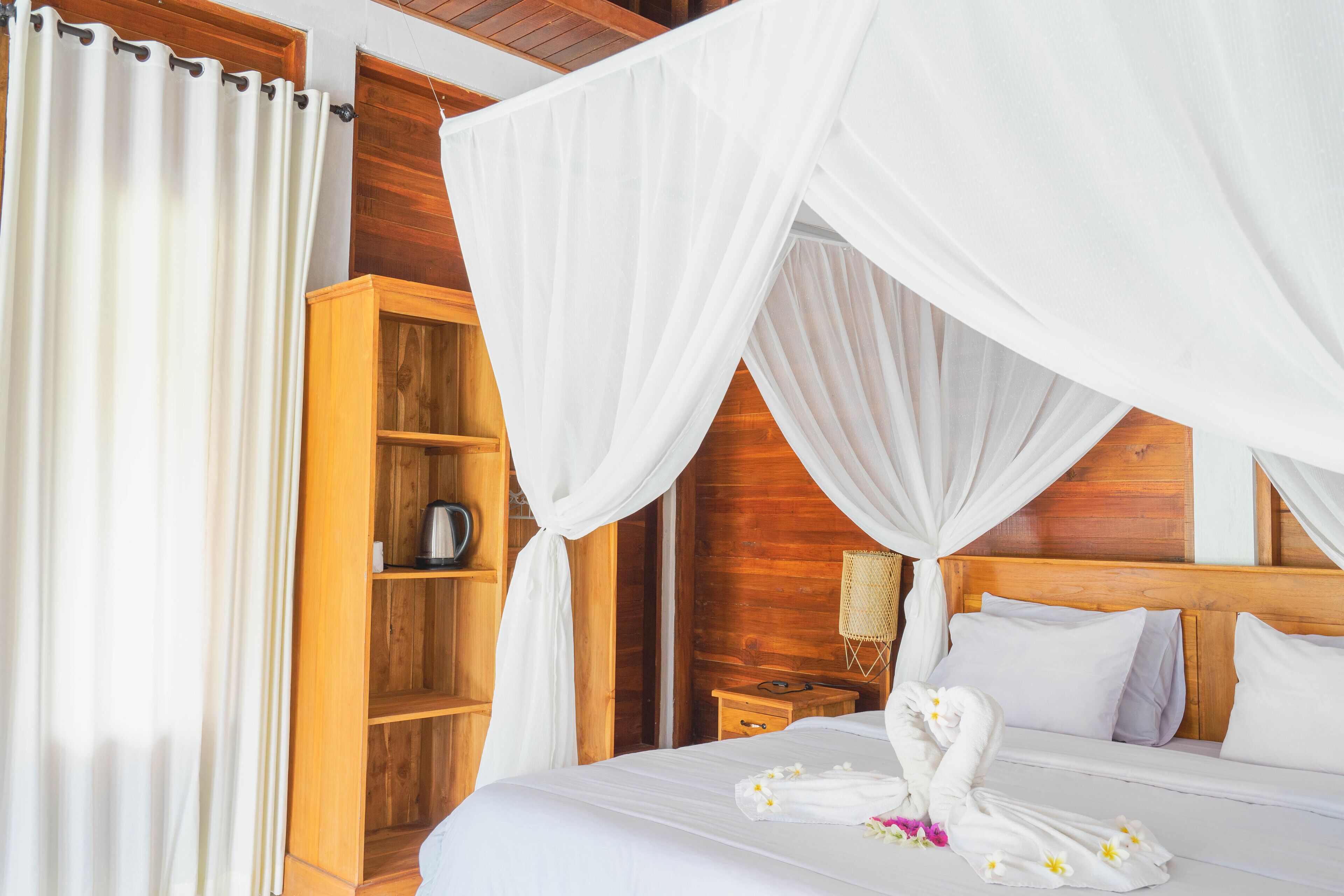 A hotel room with a wooden headboard, a canopy bed, a wooden wardrobe, and a bedside table with a lamp.