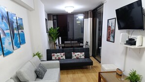 Una sala de estar con un sofá gris, un sofá negro con cojines estampados, una mesa de centro de madera y un televisor en la pared.