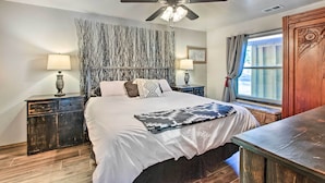 Un dormitorio con una cama grande, dos mesitas de noche con lámparas, un ventilador de techo, una ventana con cortinas y un panel decorativo en la pared.