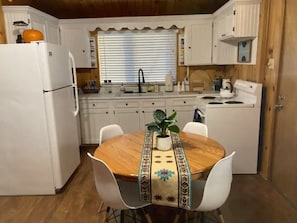 Uma cozinha compacta com eletrodomésticos brancos, uma mesa de jantar de madeira com um jogo americano estampado e uma planta em vaso.