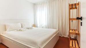 Un dormitorio con cama, cortinas blancas, estante de madera y mesita de noche.