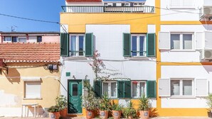 Un edificio de varios pisos con fachada amarilla y blanca, contraventanas verdes y plantas en macetas al frente.