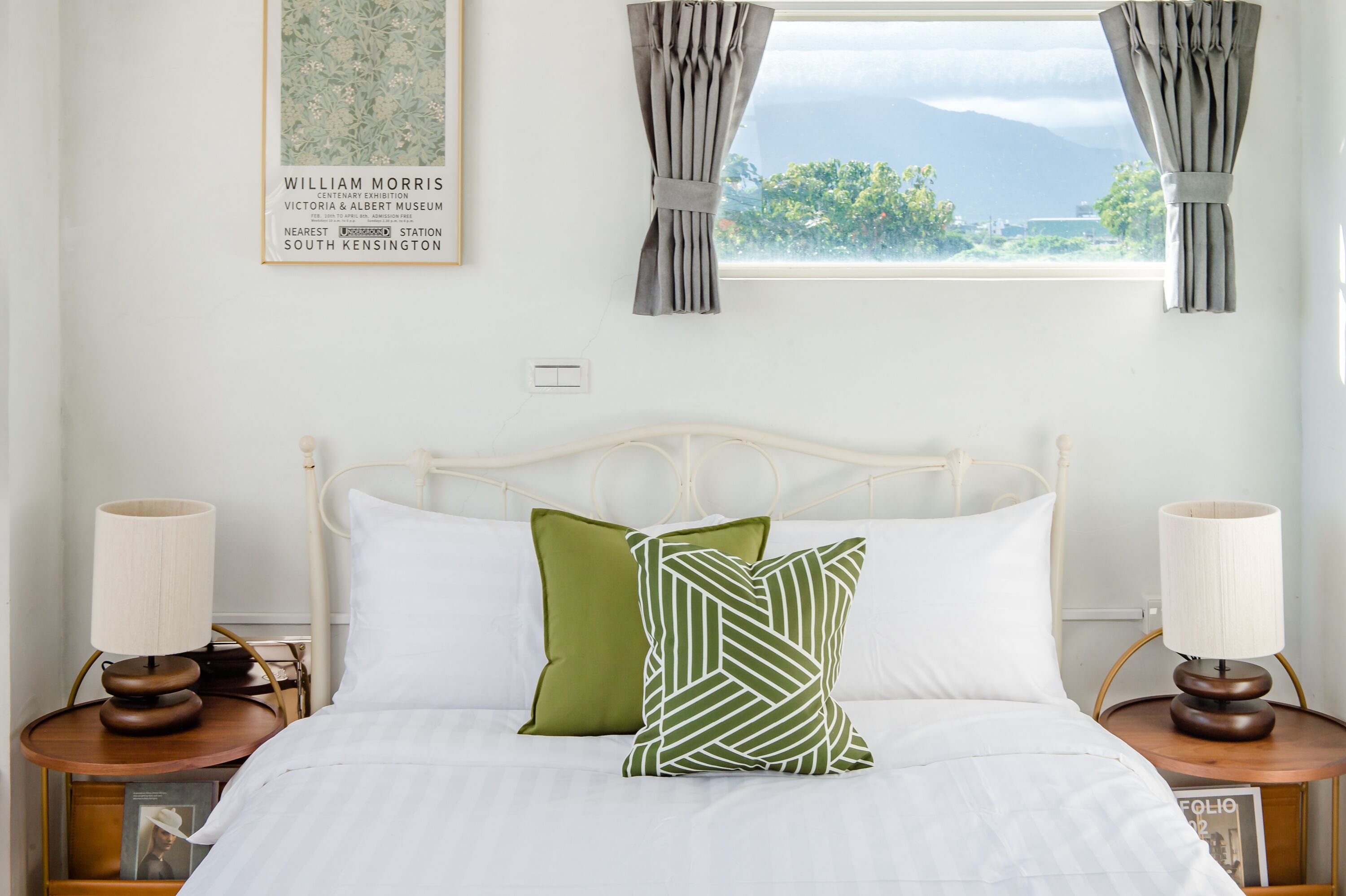 A neatly made bed with white and green pillows, a white headboard, and two bedside tables with lamps. A framed picture hangs on the wall above the bed.