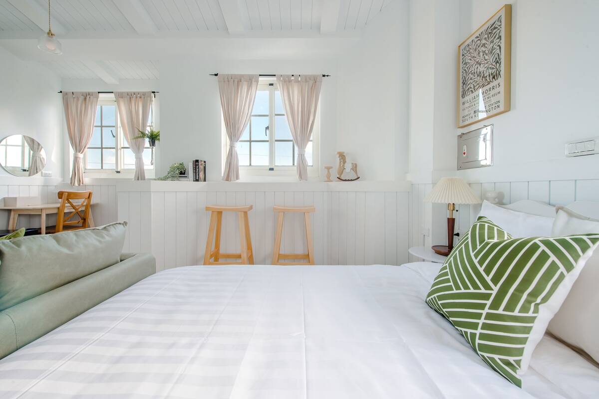 Une chambre avec un grand lit, deux tabourets en bois, une petite table, une chaise, une lampe et un tableau encadré au mur.