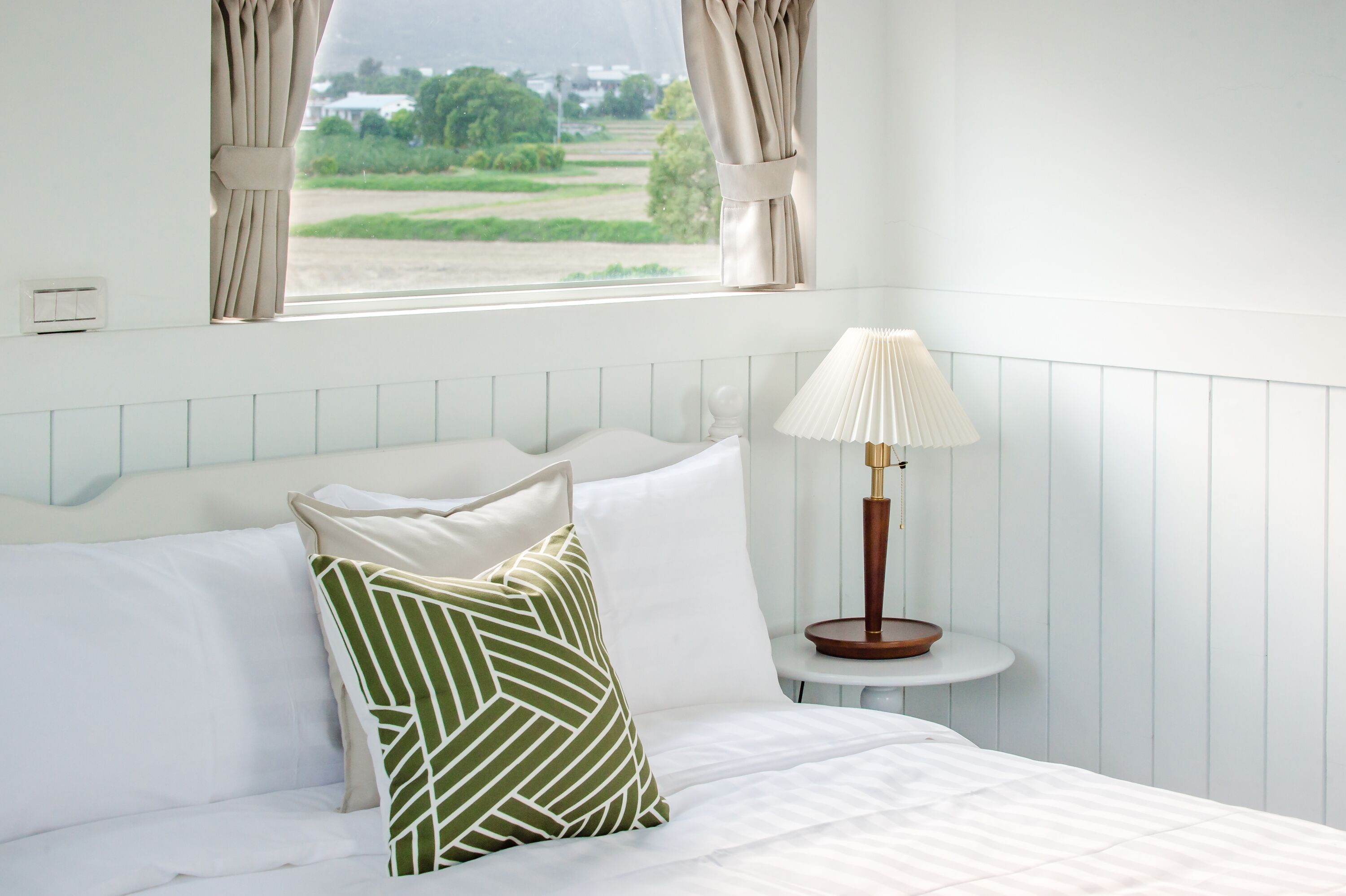 A bedroom with a bed, pillows, a bedside table with a lamp, and a window with curtains.