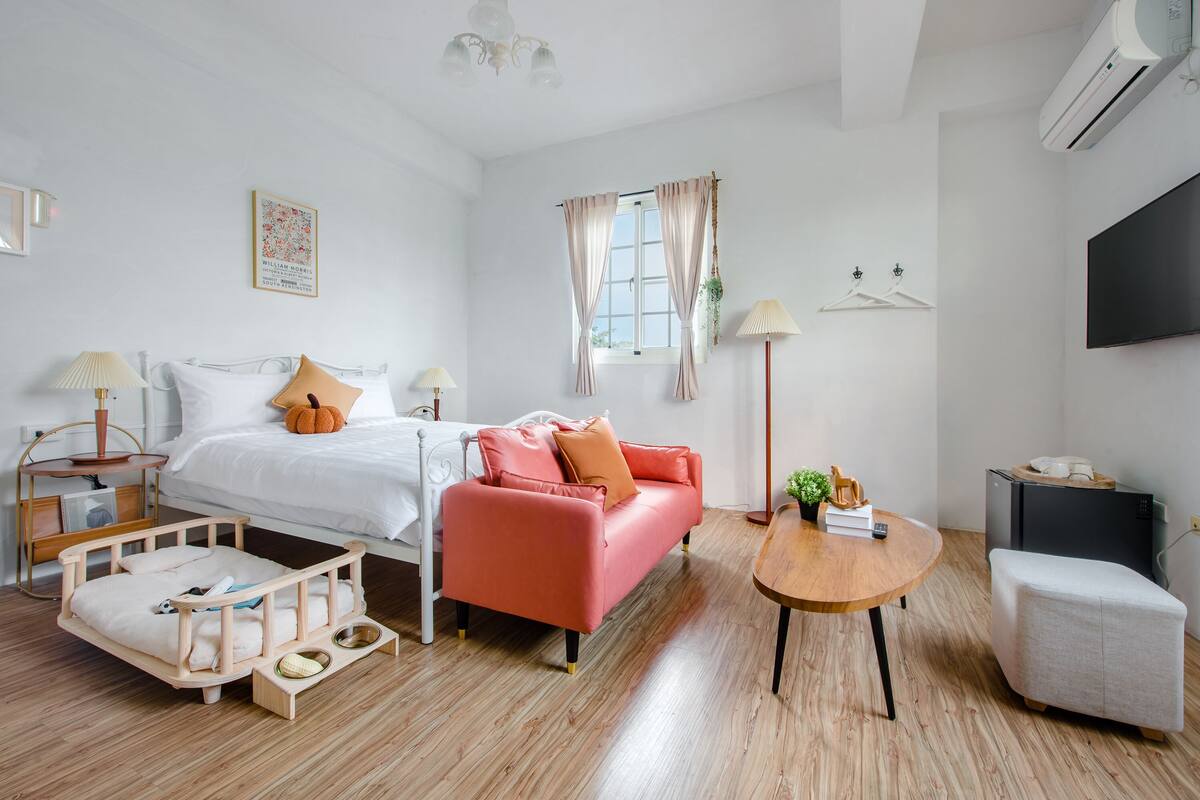 Une chambre moderne avec un lit, un canapé rose, une table basse en bois, un petit pouf blanc et un téléviseur fixé au mur.