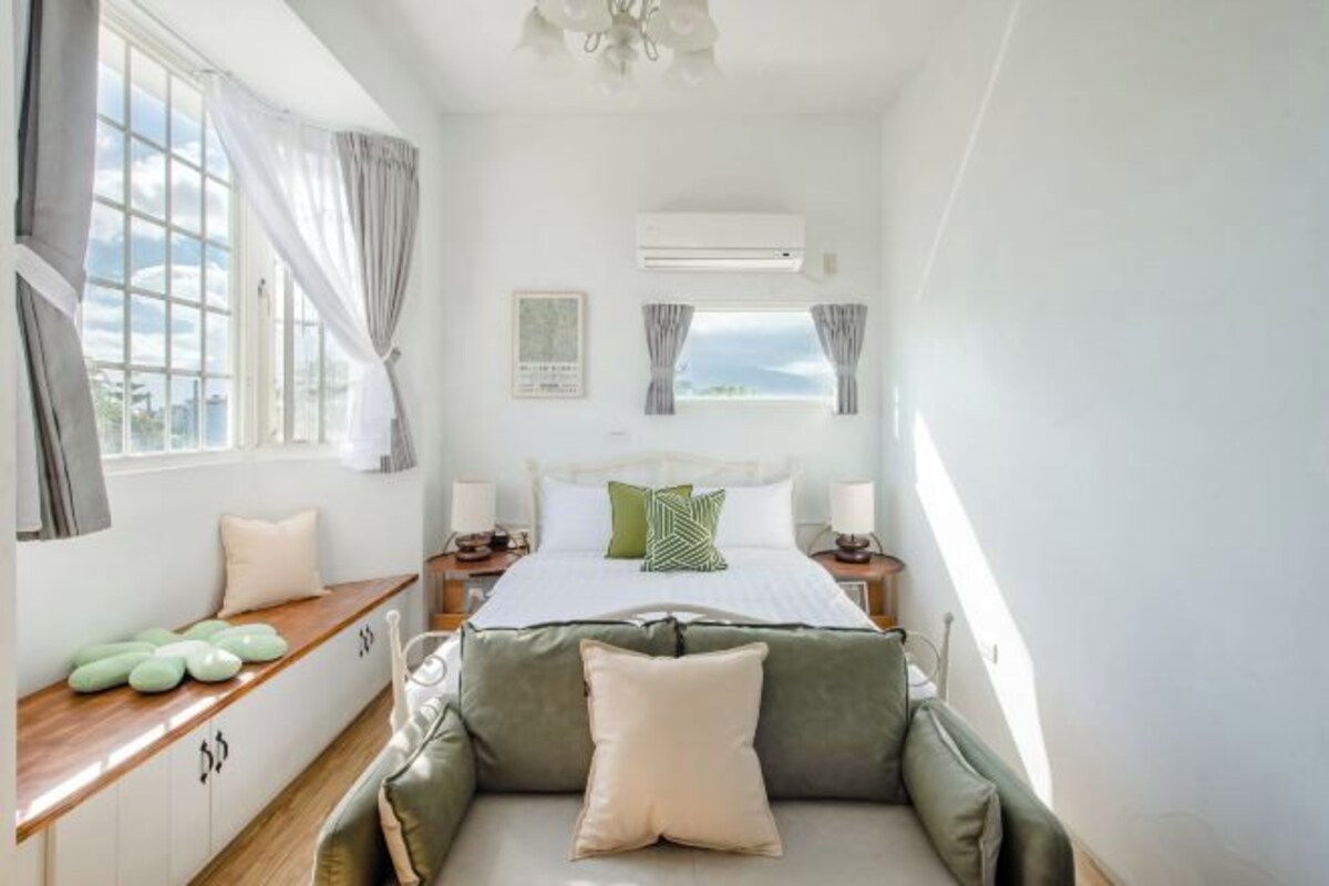 Quarto com cama, mesas de cabeceira, janela com cortinas, ar condicionado e vista para o exterior.