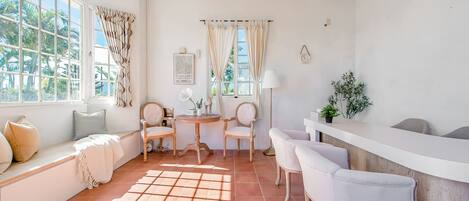 A bright room with a large window, a wooden ceiling, a round table, and a seating area with chairs and a bench.