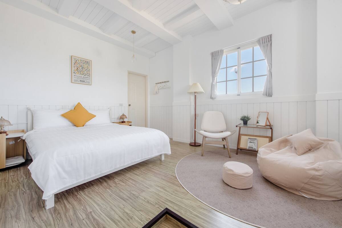 Une chambre avec un grand lit, une chaise, un fauteuil poire, une table de chevet, une lampe et une fenêtre avec des rideaux.