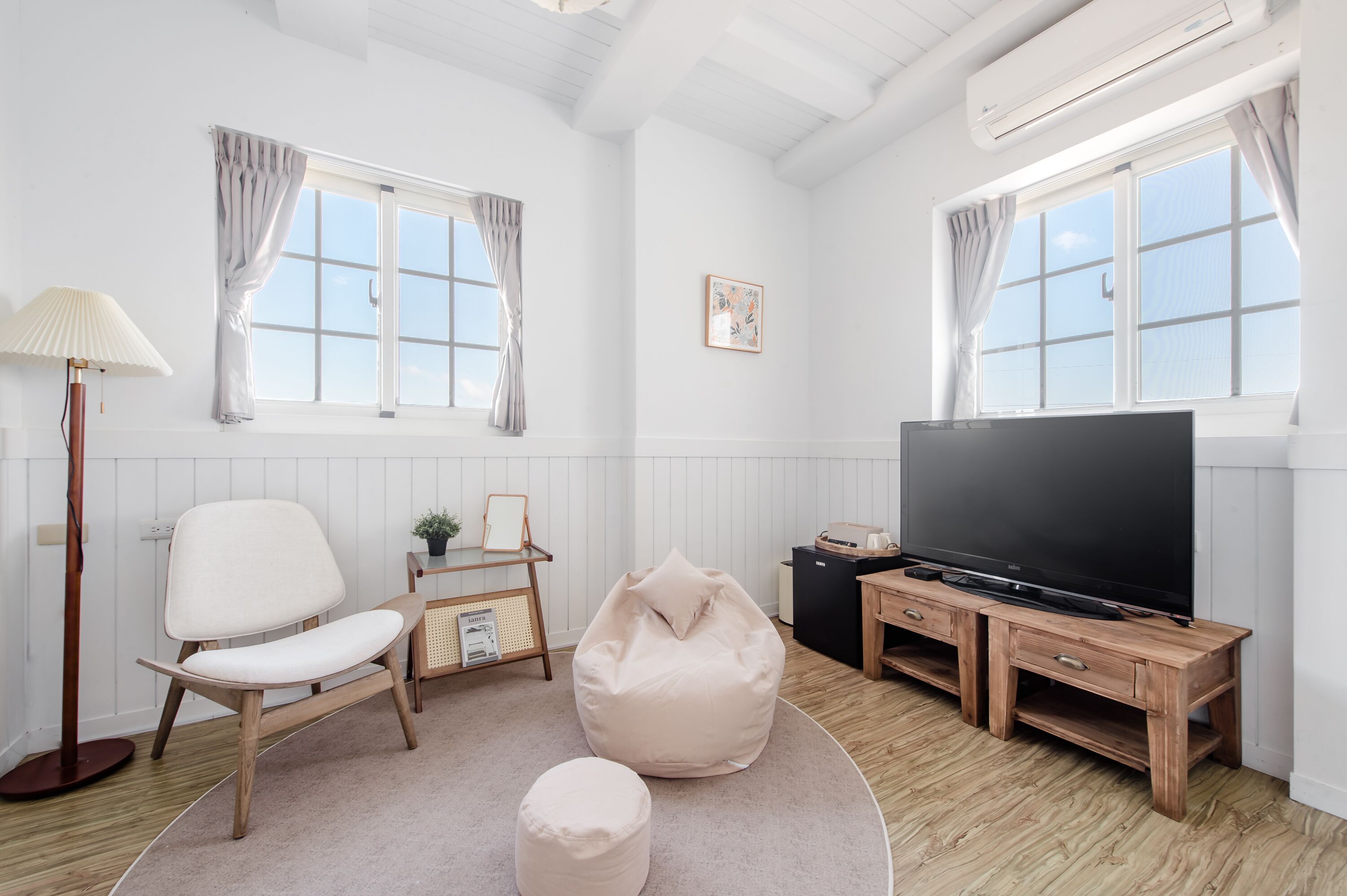 A modern living room with a white chair, a bean bag, a TV on a wooden stand, and a small table with a lamp.
