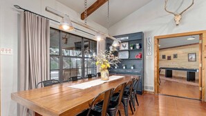 Un área de comedor con una mesa de madera, sillas negras y un jarrón con flores. Hay ventanas con cortinas, una estantería con plantas y un letrero que dice