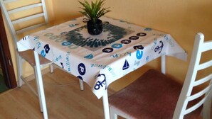 Une petite table à manger recouverte d’une nappe, deux chaises blanches, une plante en pot et une étagère ornée d’objets décoratifs.