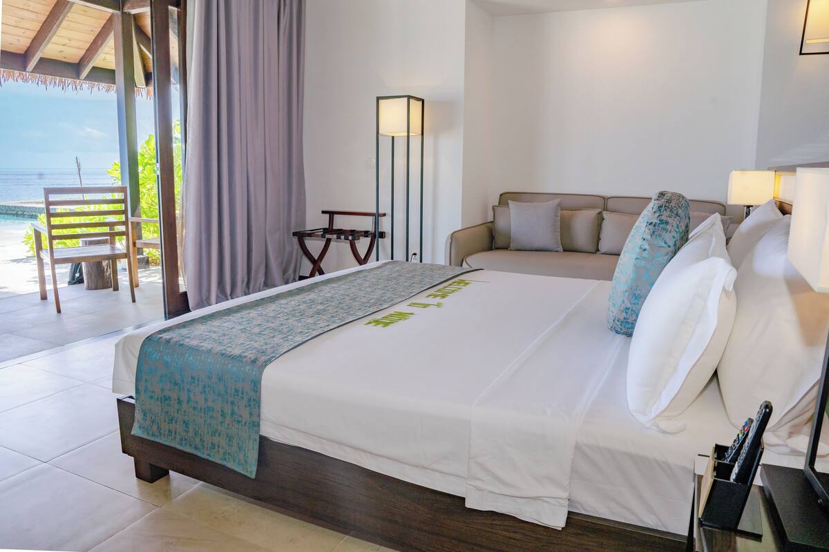 Habitación de hotel con cama, sofá, lámpara de pie y vistas a la playa.