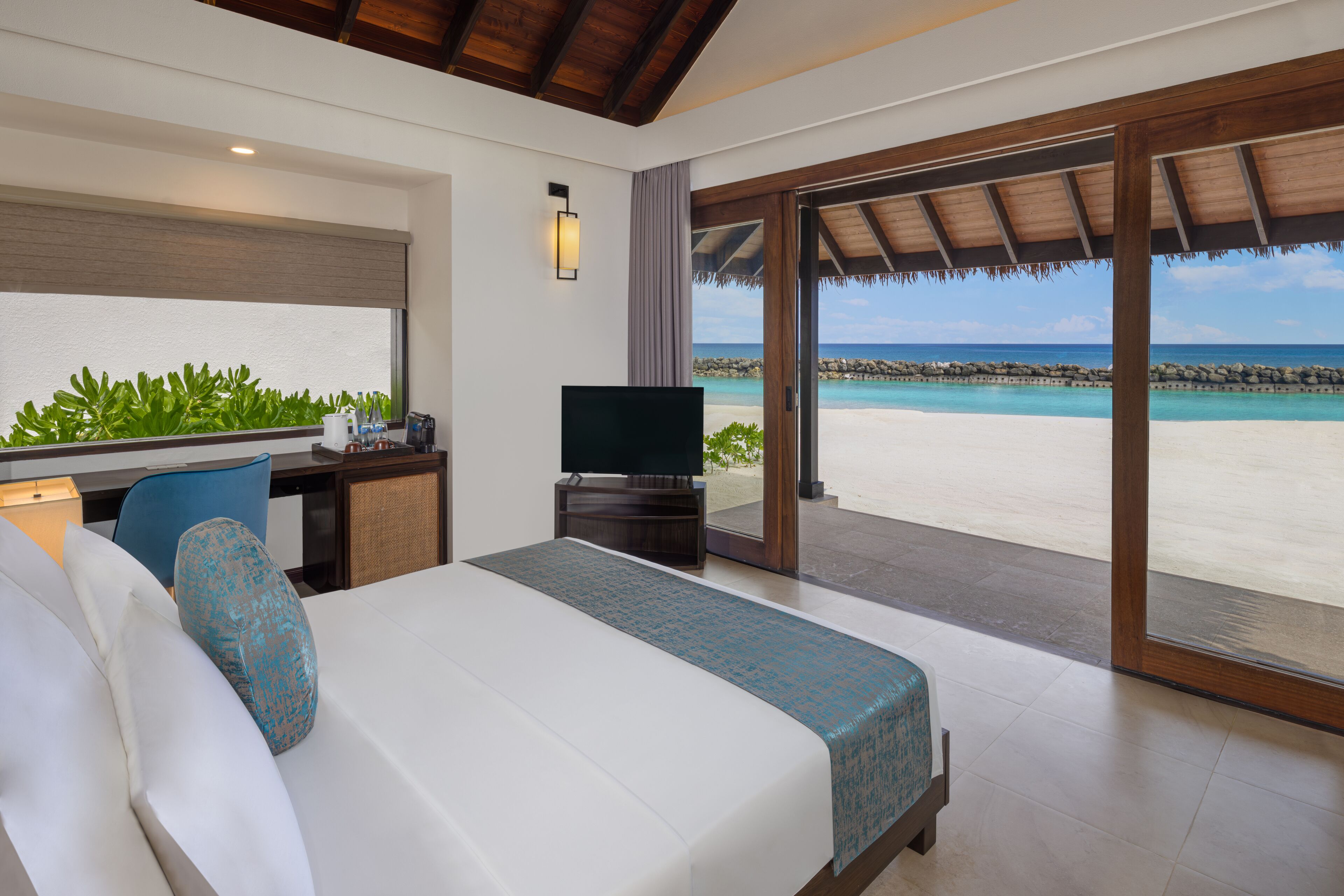 A hotel room with a bed, a desk, a television, and a large window with a view of a beach and ocean.