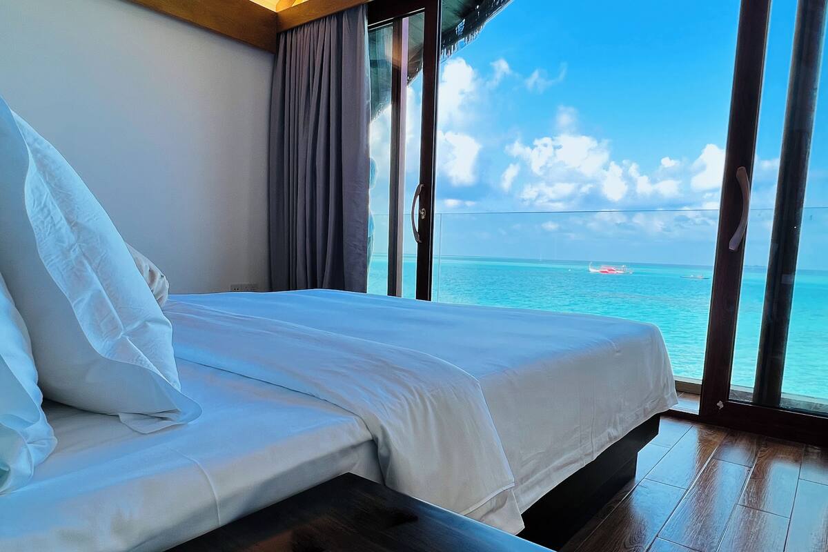 Un dormitorio con cama, mesita de noche de madera y un ventanal con vista al océano y barcos.