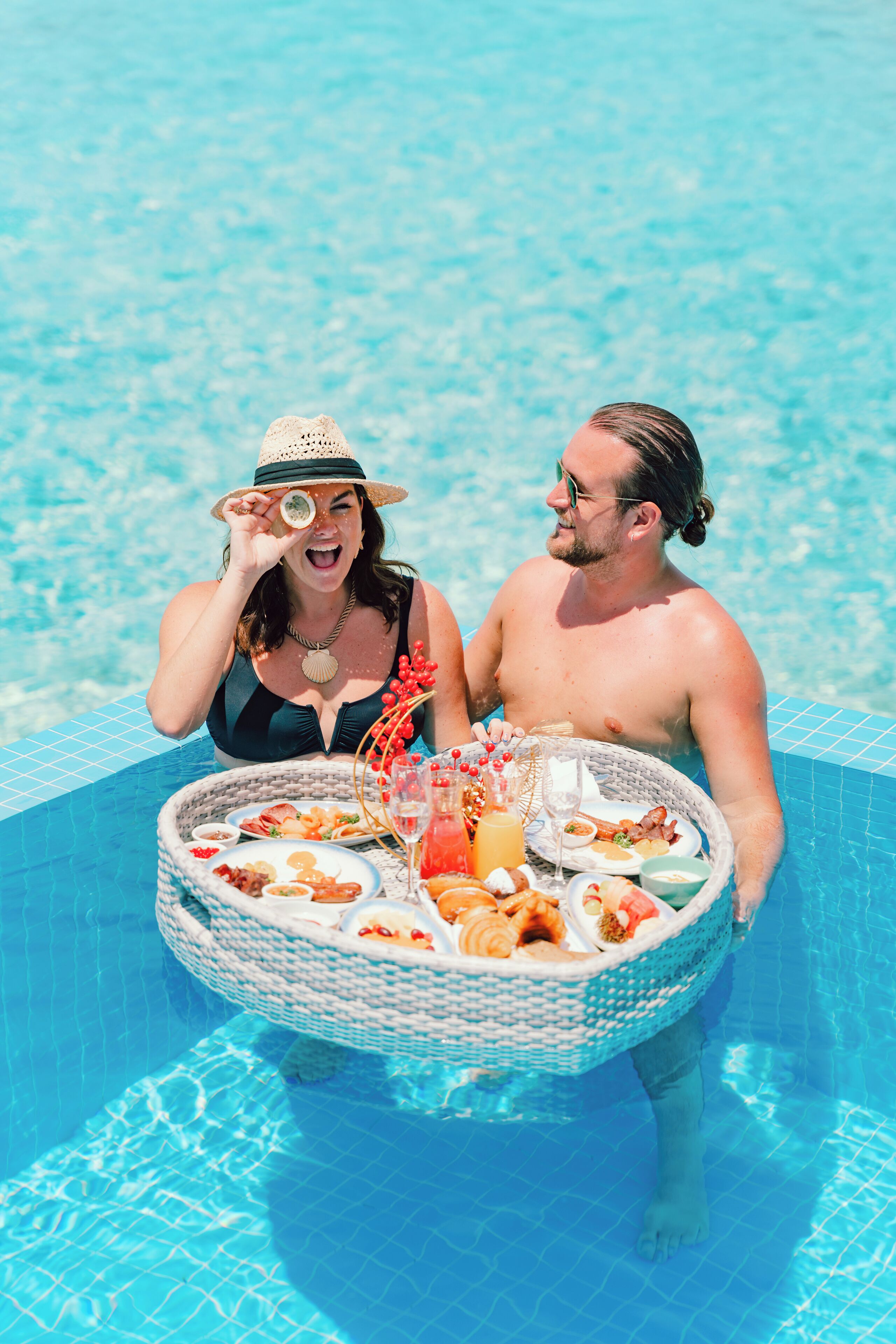 A couple in a pool with a wicker basket containing food and drinks.