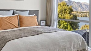 A bed with a grey headboard and beige bedding, a nightstand with a framed picture, and a view of mountains and trees through a window. - Ultimate Views - Tikumu Lake House (Queenstown)
