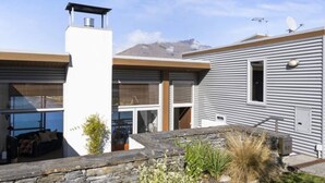 A modern house with a stone retaining wall, a covered outdoor seating area, and a mountain in the background. - Ultimate Views - Tikumu Lake House (Queenstown)