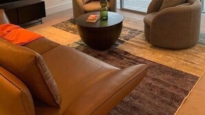 A modern living room with a brown leather sofa, two armchairs, a coffee table, and a TV. A large window offers a view of a body of water and mountains. - Ultimate Views - Tikumu Lake House (Queenstown)