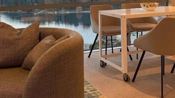 A modern living room with a large window overlooking a lake and mountains. There is a sofa, a dining table with chairs, and a small cart. - Ultimate Views - Tikumu Lake House (Queenstown)