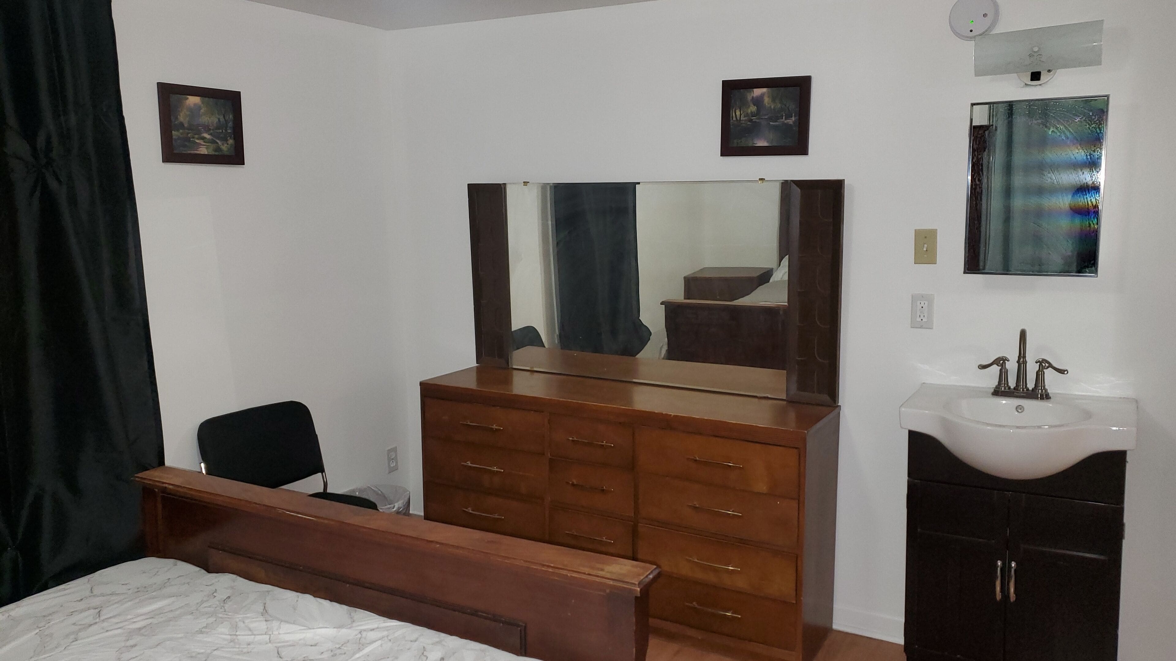 Een slaapkamer met een houten dressoir, een grote spiegel, een nachtkastje, een stoel en een witte wastafel.