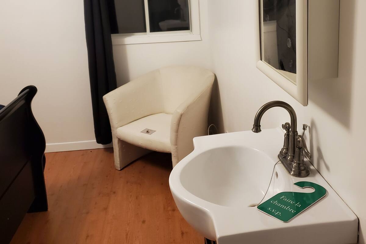 Une petite salle de bain avec un lavabo blanc, un miroir, une chaise et une fenêtre avec un rideau noir.