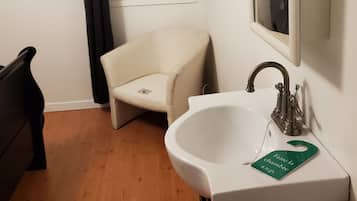 Een compacte badkamer met een wit wastafelmeubel, een spiegel, een stoel en een raam met een zwart gordijn.