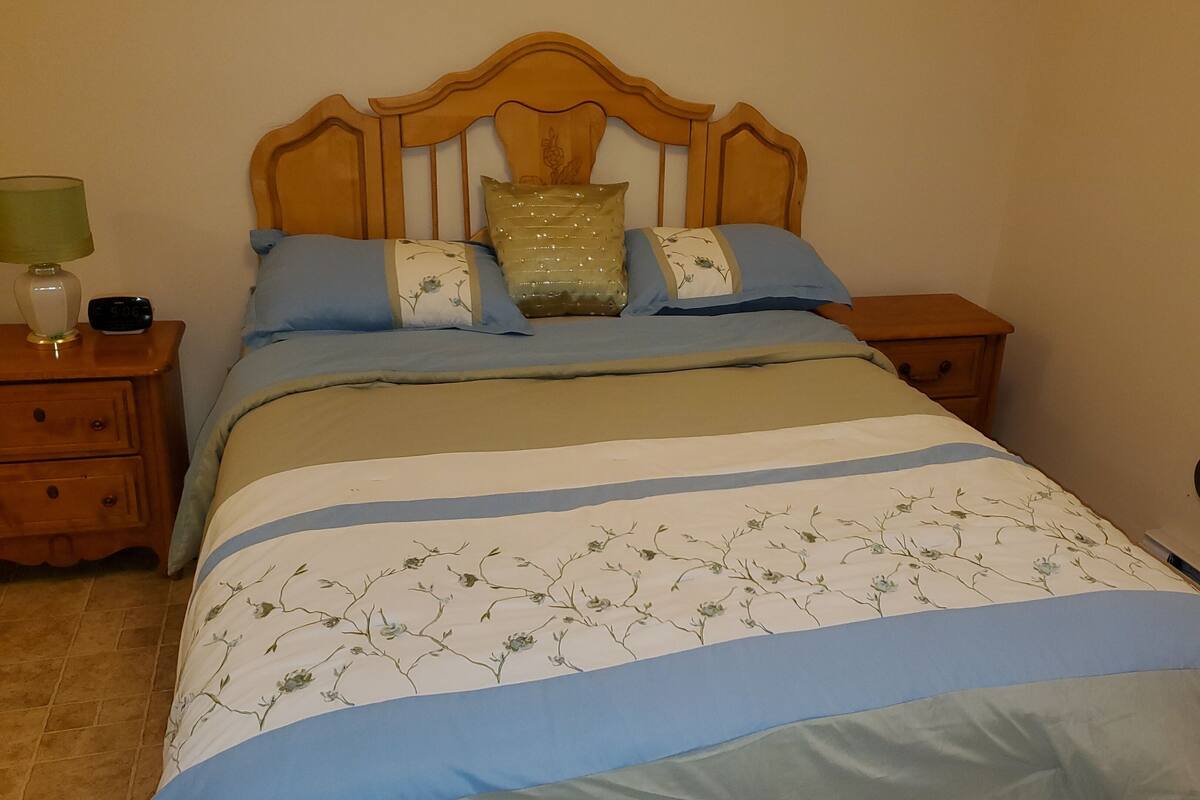 Un lit avec une couvre-lit à motifs floraux, deux oreillers, une tête de lit en bois et une table de chevet avec une lampe et une horloge.