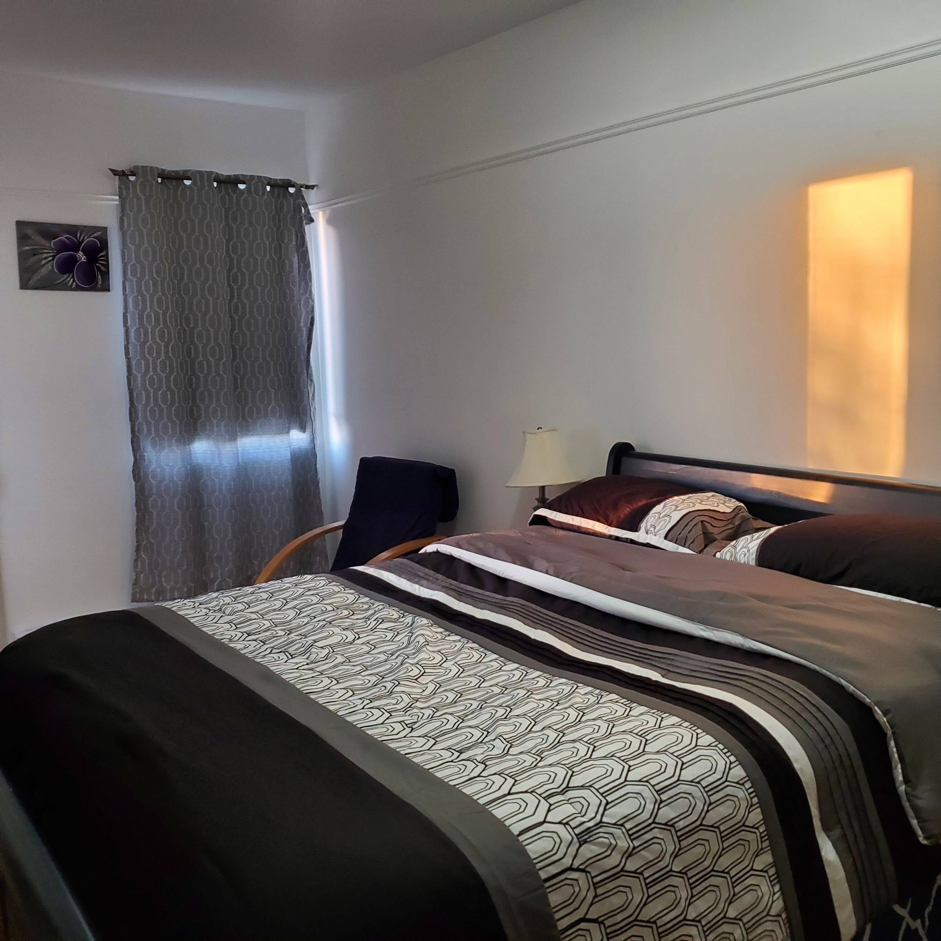 Una cama bien hecha con ropa de cama estampada, una silla, una lámpara y una ventana con cortinas.