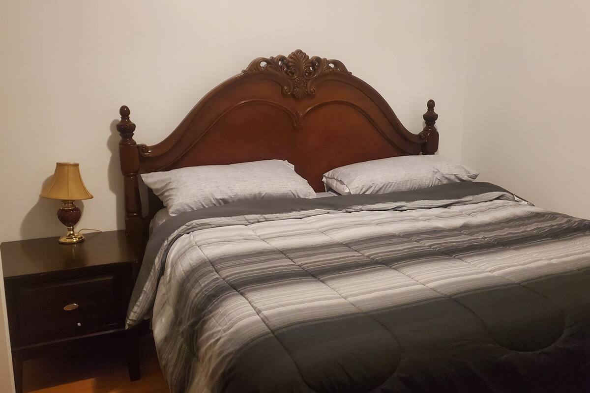 Un lit bien fait, avec une couette à rayures et deux oreillers, une table de chevet en bois avec une lampe, et une porte sur le côté gauche.