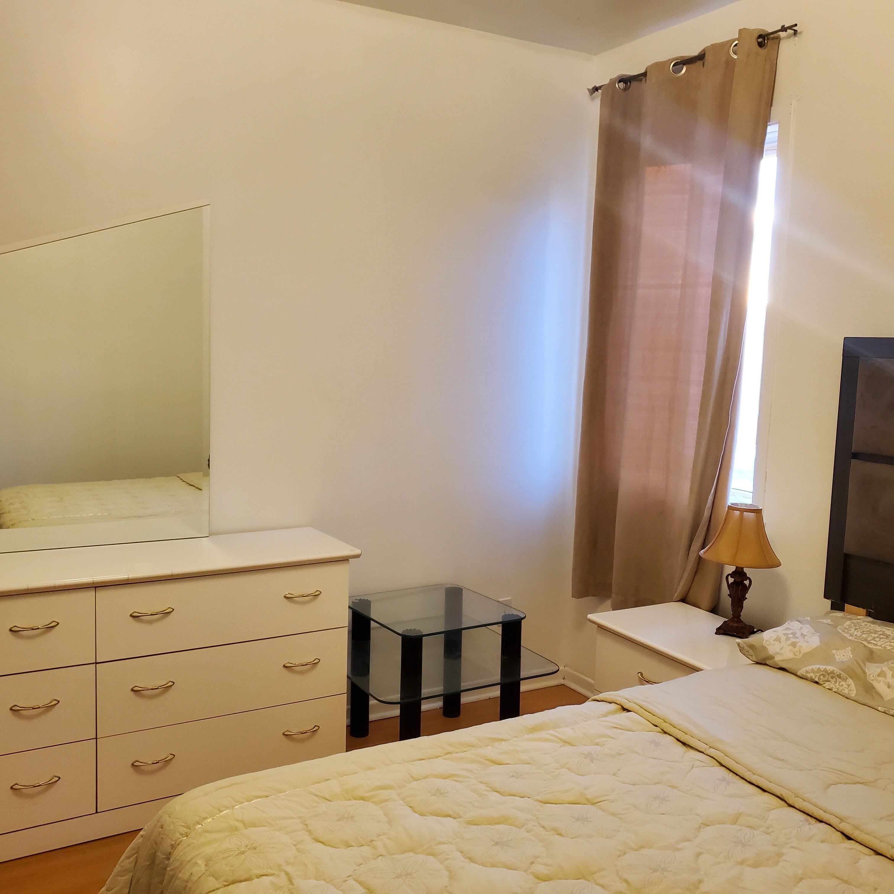 Une chambre à coucher comprenant un lit, une commode avec des tiroirs, une petite table, une lampe et une fenêtre avec des rideaux.