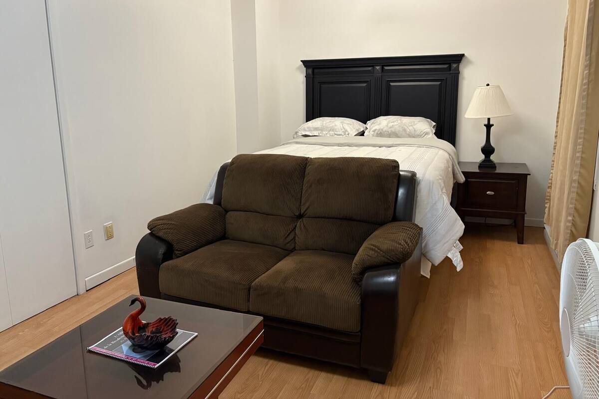 Une petite pièce, bien rangée, avec un lit, une chaise, une table de chevet avec une lampe et un ventilateur.